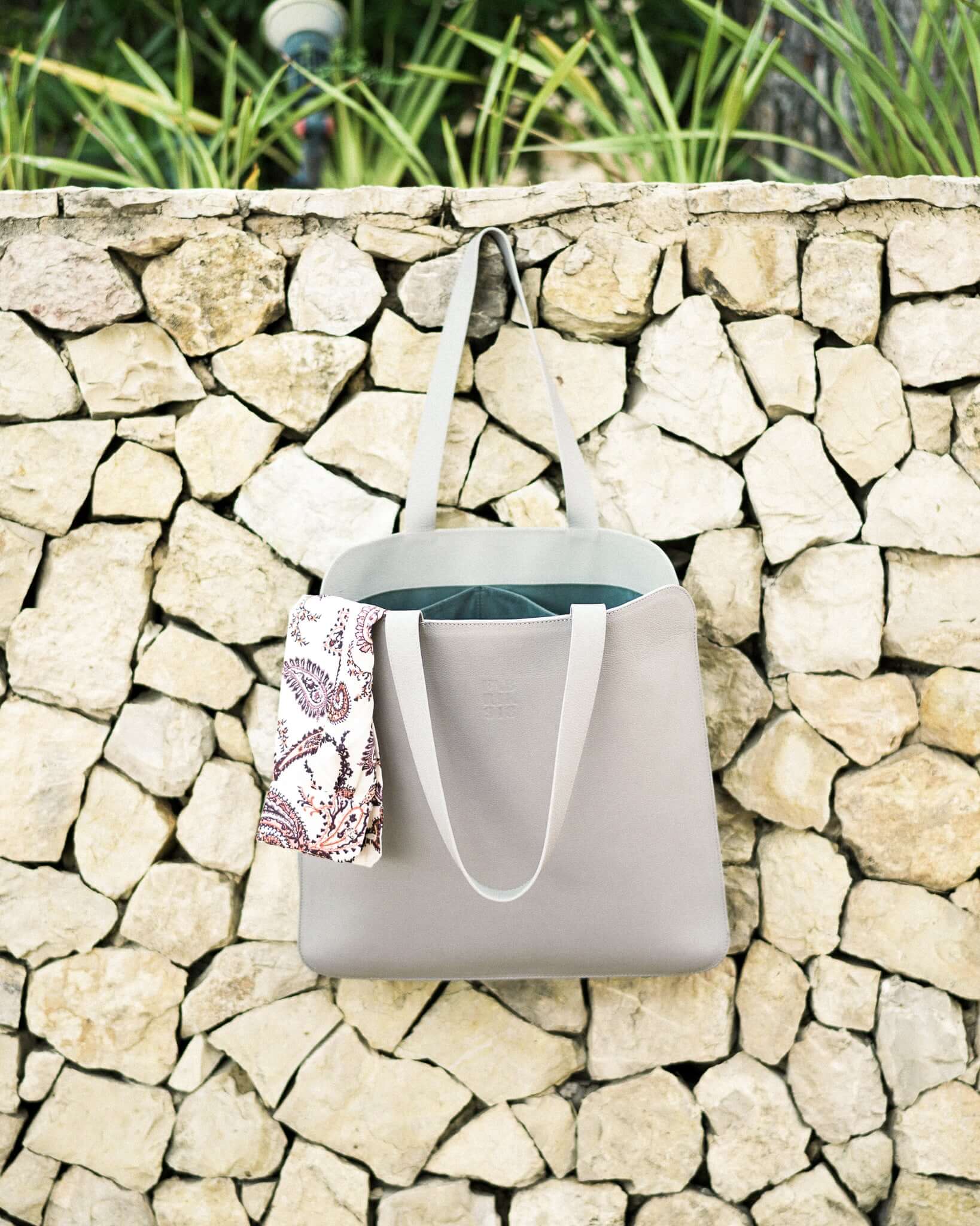 Handcrafted leather tote bag hanging on stone wall with a patterned scarf peeking out, showcasing durability and elegance.